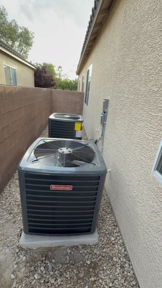 Two HVAC outdoor condenser units installed on gravel along the beige stucco side wall of a residential house.