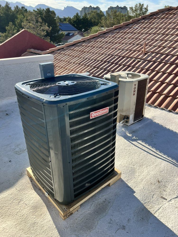 A dark green Goodman air conditioning unit sits on a wooden pallet on a flat rooftop, next to an older, tan unit.