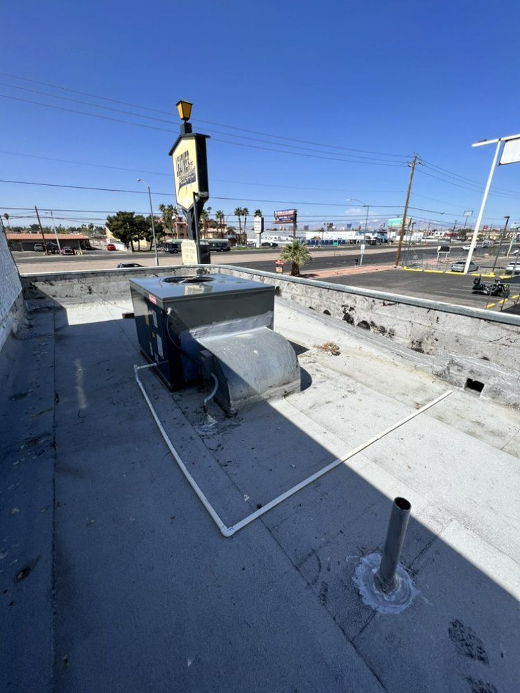 A flat rooftop with a rectangular HVAC unit, a metal pipe, and a white conduit line, under a clear blue sky.