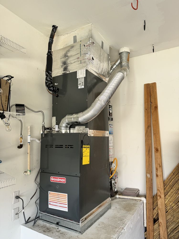 A Goodman HVAC furnace unit standing on a raised concrete base in a garage, connected to metal ductwork and piping.