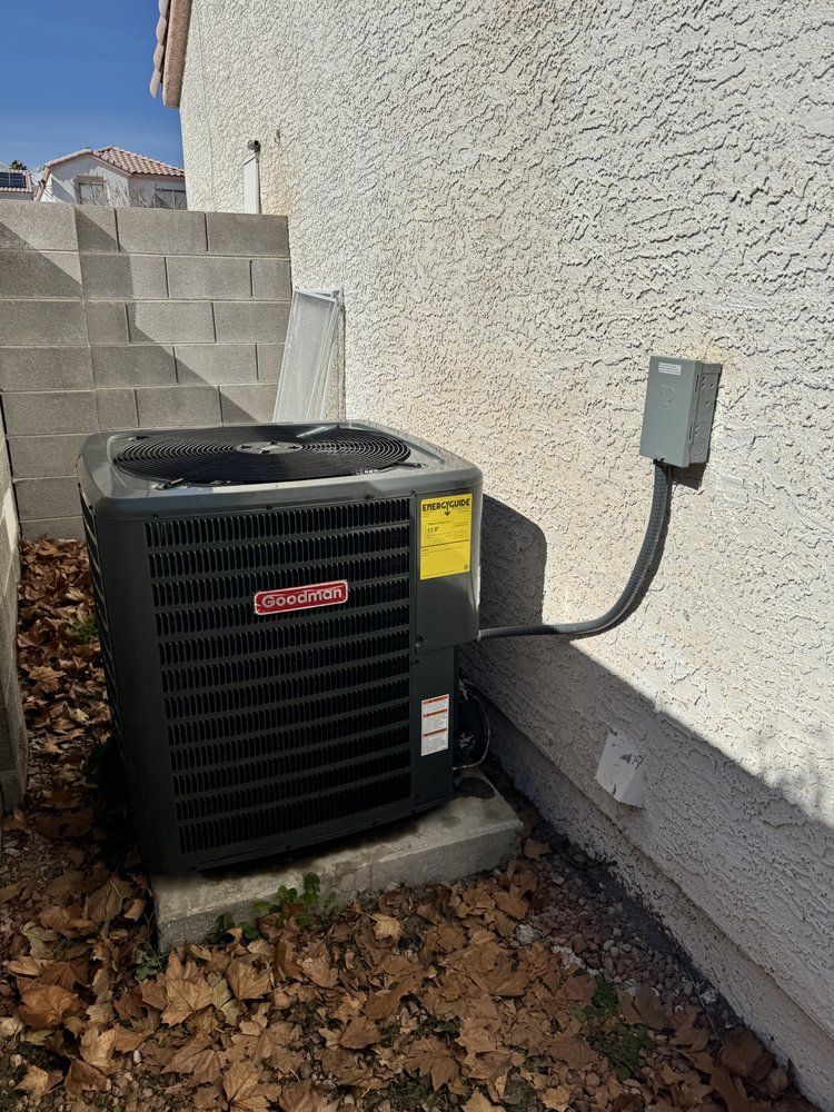 A gray outdoor Goodman air conditioning unit sits on a concrete pad next to a stucco house wall with an electrical box.