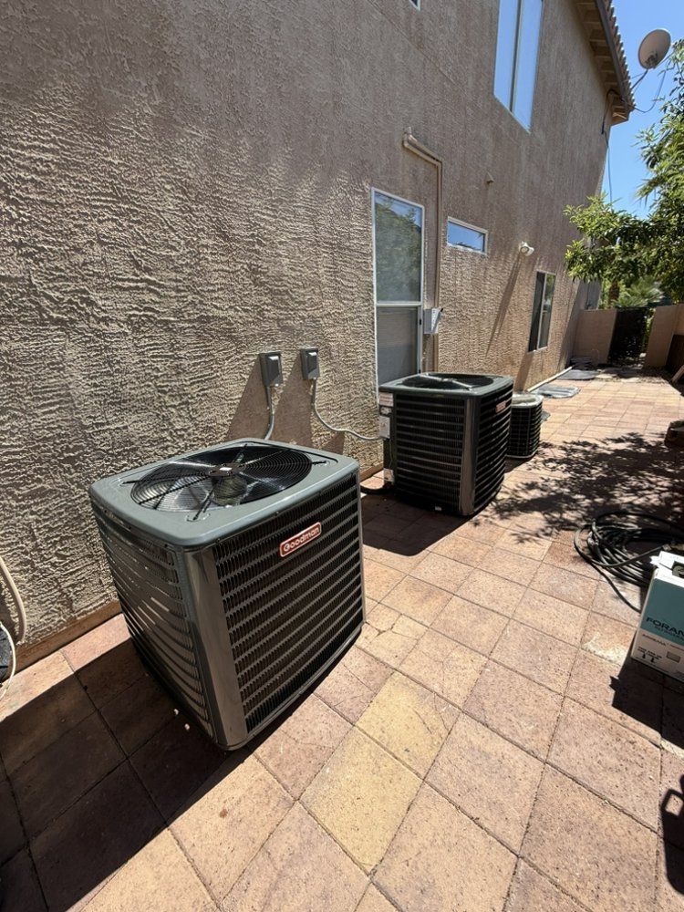 Two gray HVAC condenser units sit on a tiled patio next to the beige stucco wall of a house on a sunny day.