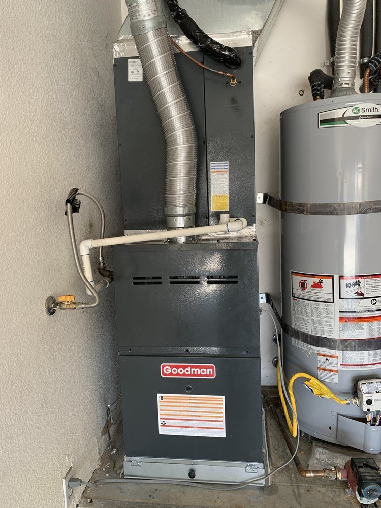 A dark grey Goodman furnace unit stands next to a cylindrical grey water heater in a utility space.
