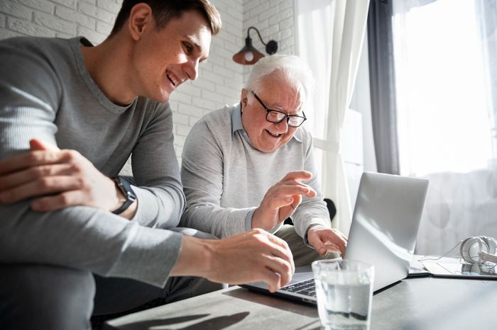 Man shows an elderly person a laptop screen, both smiling, indoor setting, sunny.