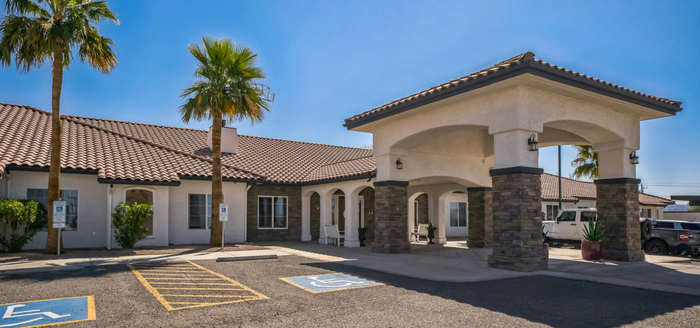 A beige, single-story building with a covered entrance; palm trees in front under a blue sky.