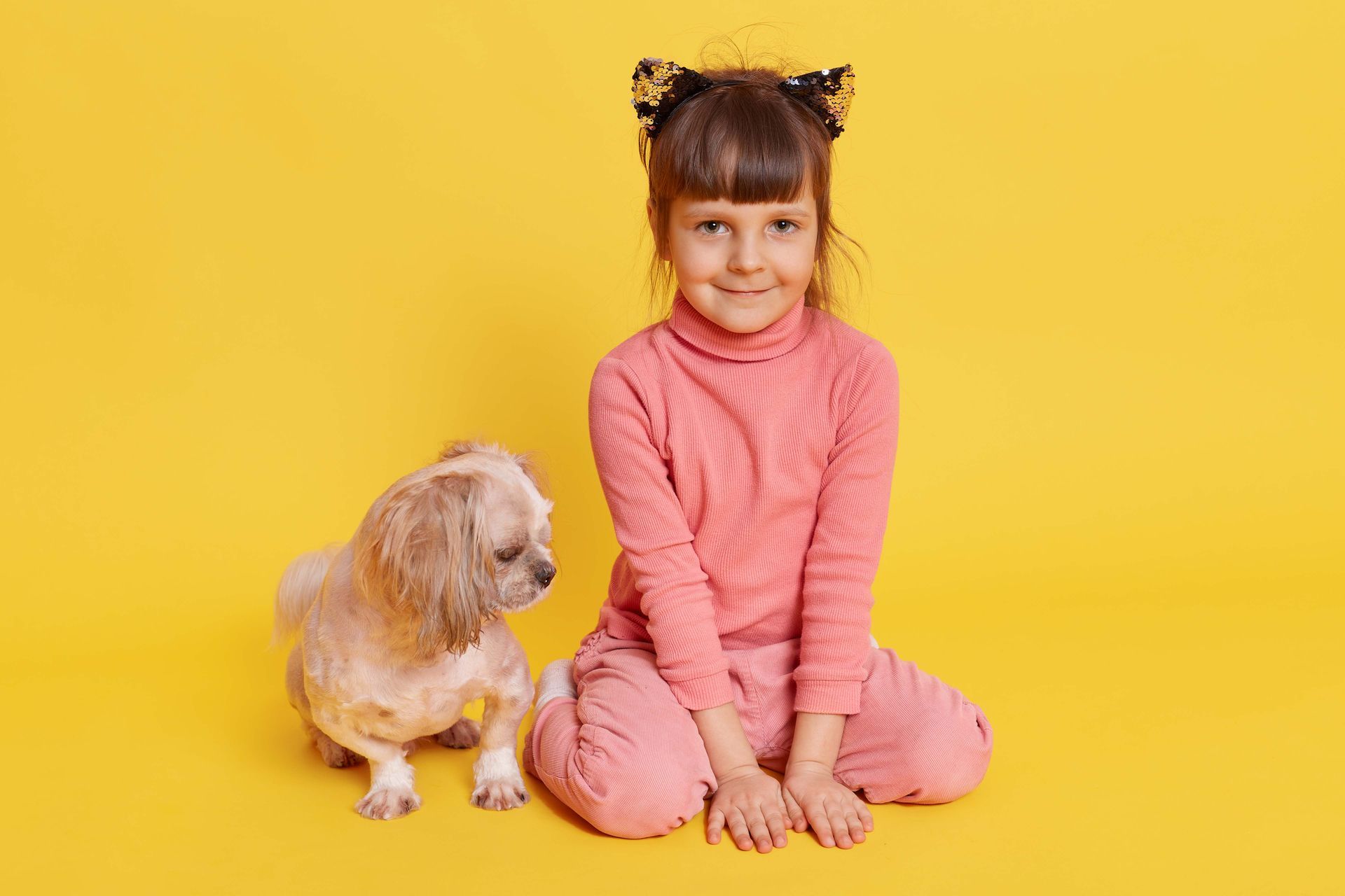 Young child in a pink outfit sitting beside a small tan dog against a yellow background