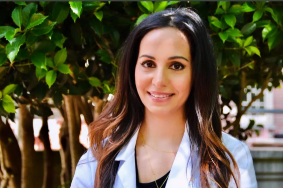 Smiling person in a white coat standing outdoors in front of green foliage.