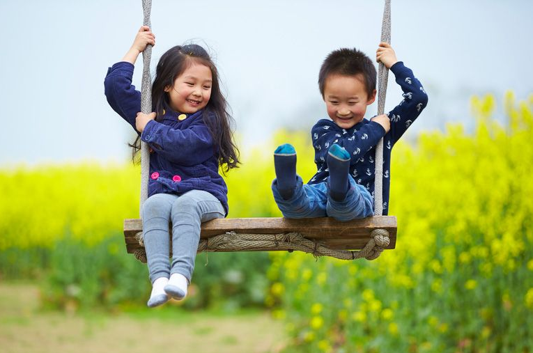 Two children on a wooden swing in a flower field, smiling and kicking their feet.