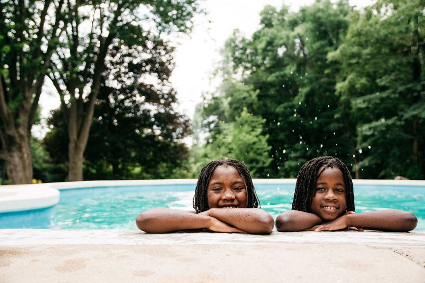 Two children resting their arms on a pool edge, smiling in front of a blue swimming pool and trees.