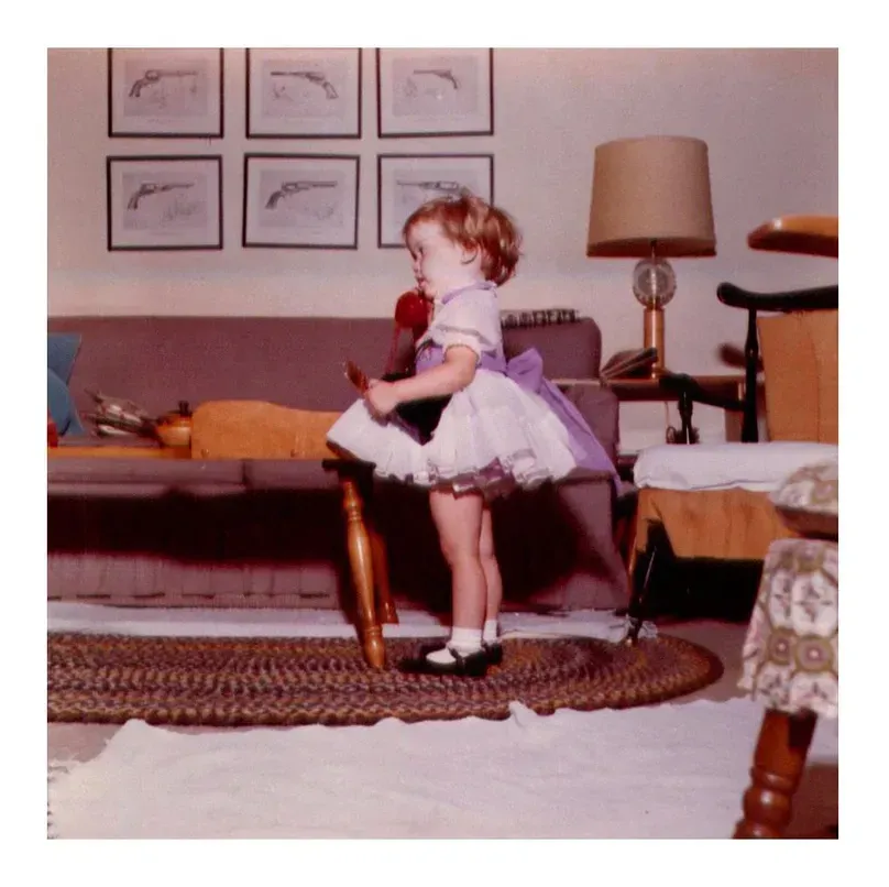 Child in a white tutu stands on a table in a living room with framed pictures and a lamp.