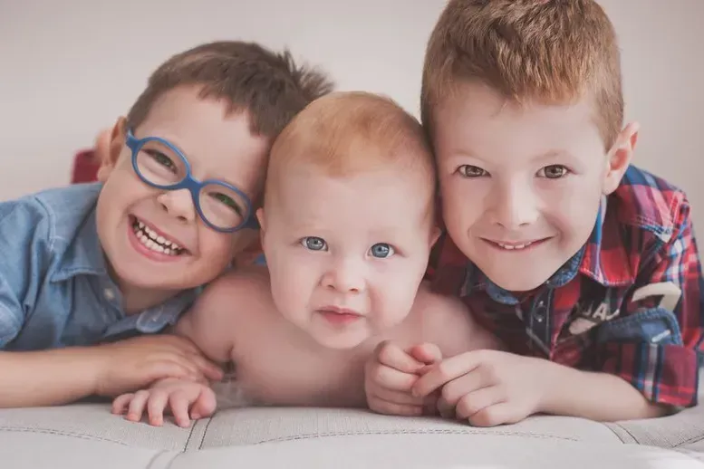 Three smiling children lying close together on a couch, one wearing blue glasses.