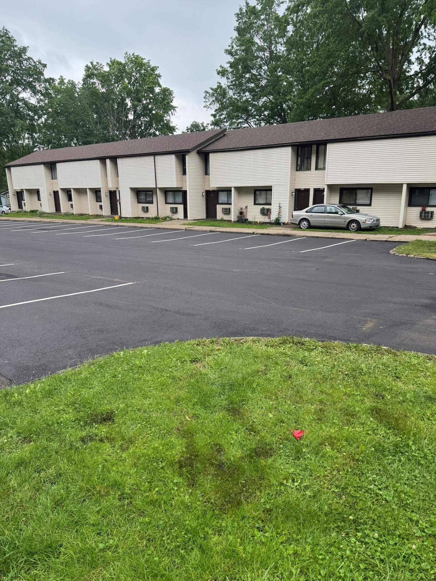 Low-rise apartment building with parked cars beside a wet asphalt lot and grassy foreground