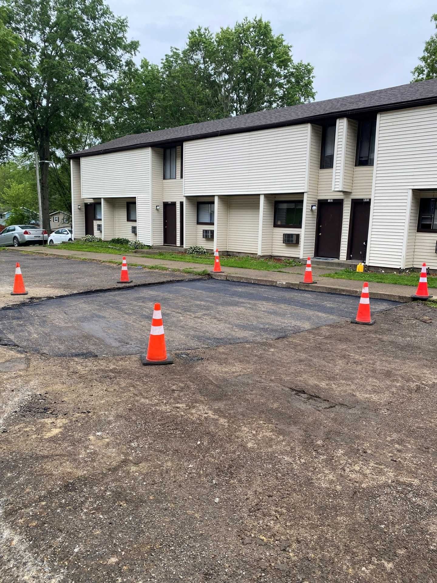 Orange cones mark a gravel parking area in front of a two-story apartment building with white siding.