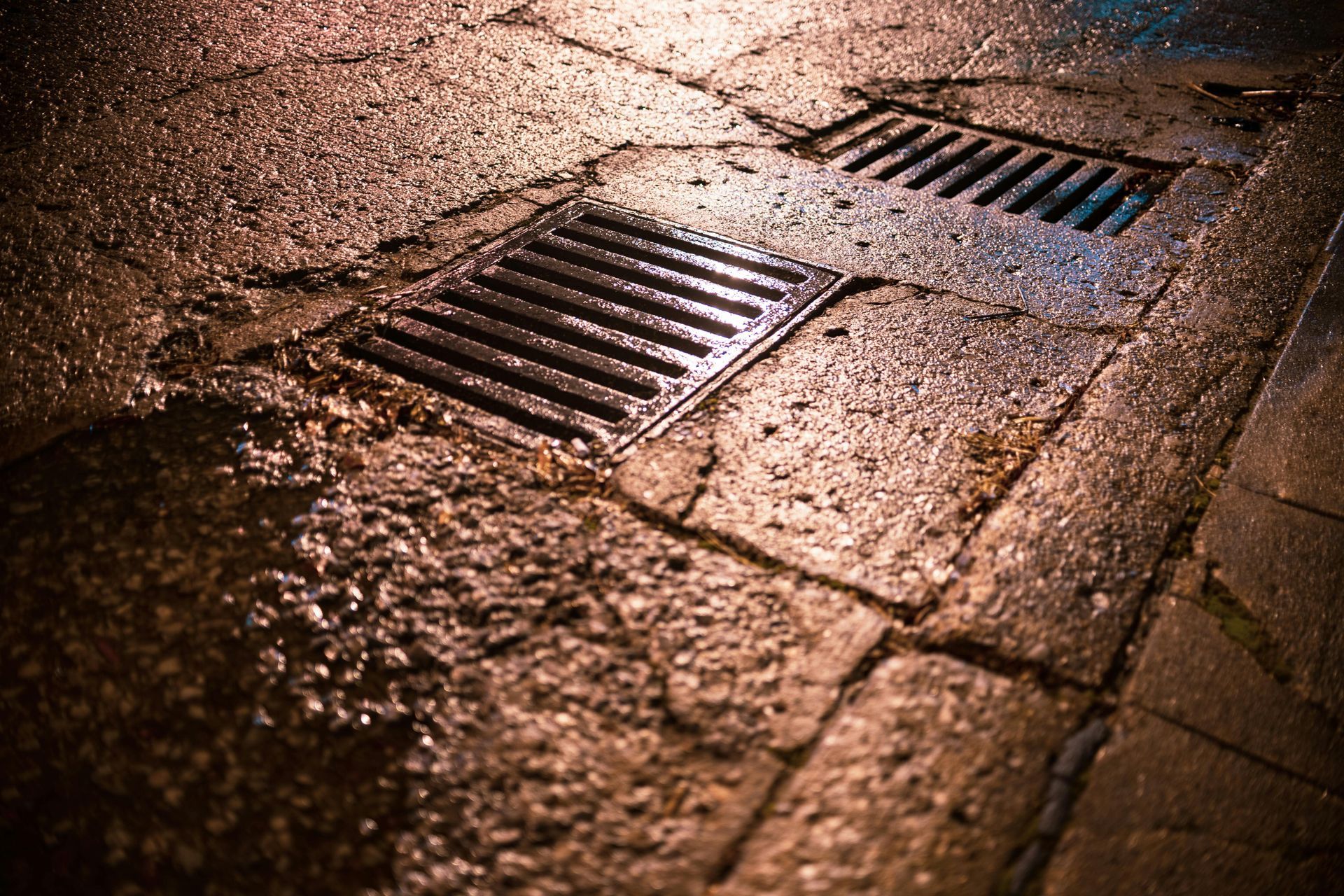 Rain-soaked cobblestone street with metal storm drains reflecting warm light