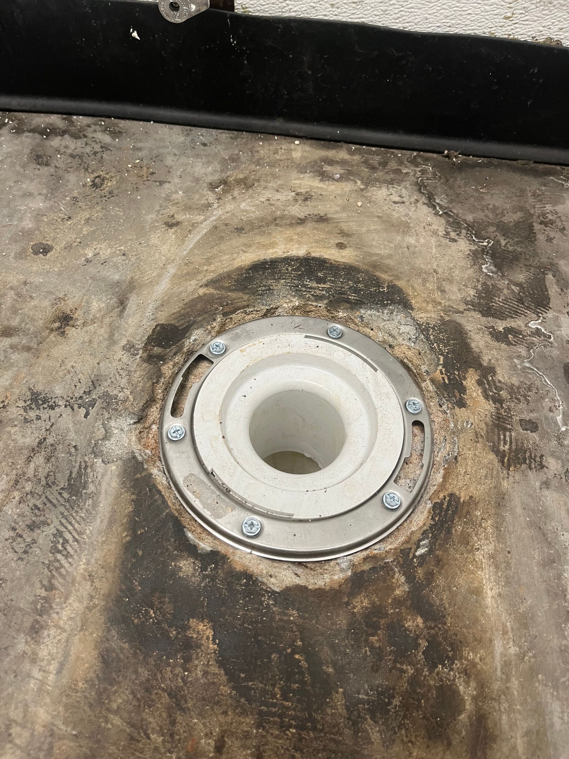 Close-up of a white toilet flange opening set in a stained concrete floor, with bolts around the rim