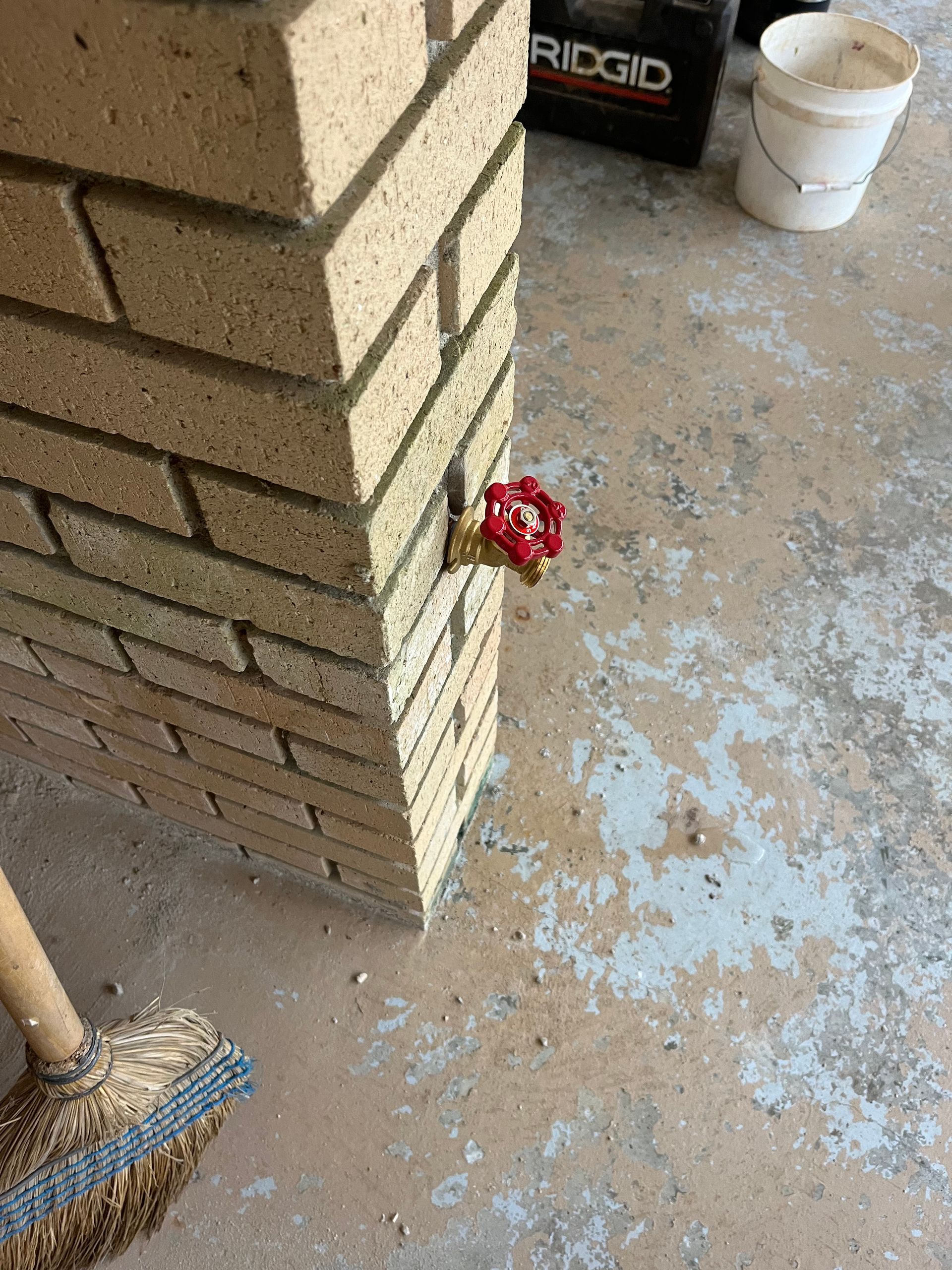 Stacked bricks beside a white bucket and broom in a garage, with a small red flower tucked into the stack.