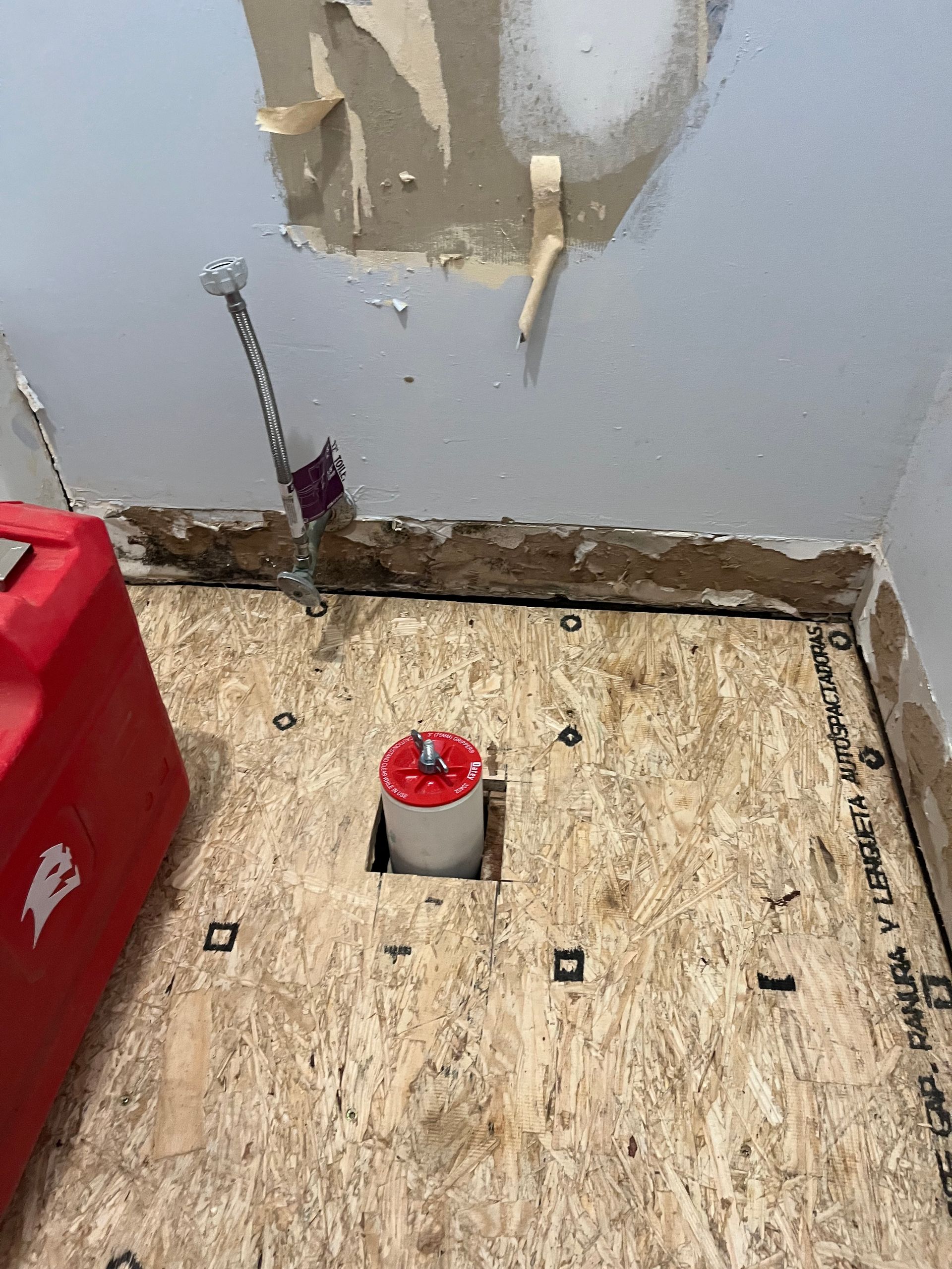 Partially stripped room corner with exposed subfloor, damaged wall, and a pipe opening in the floor