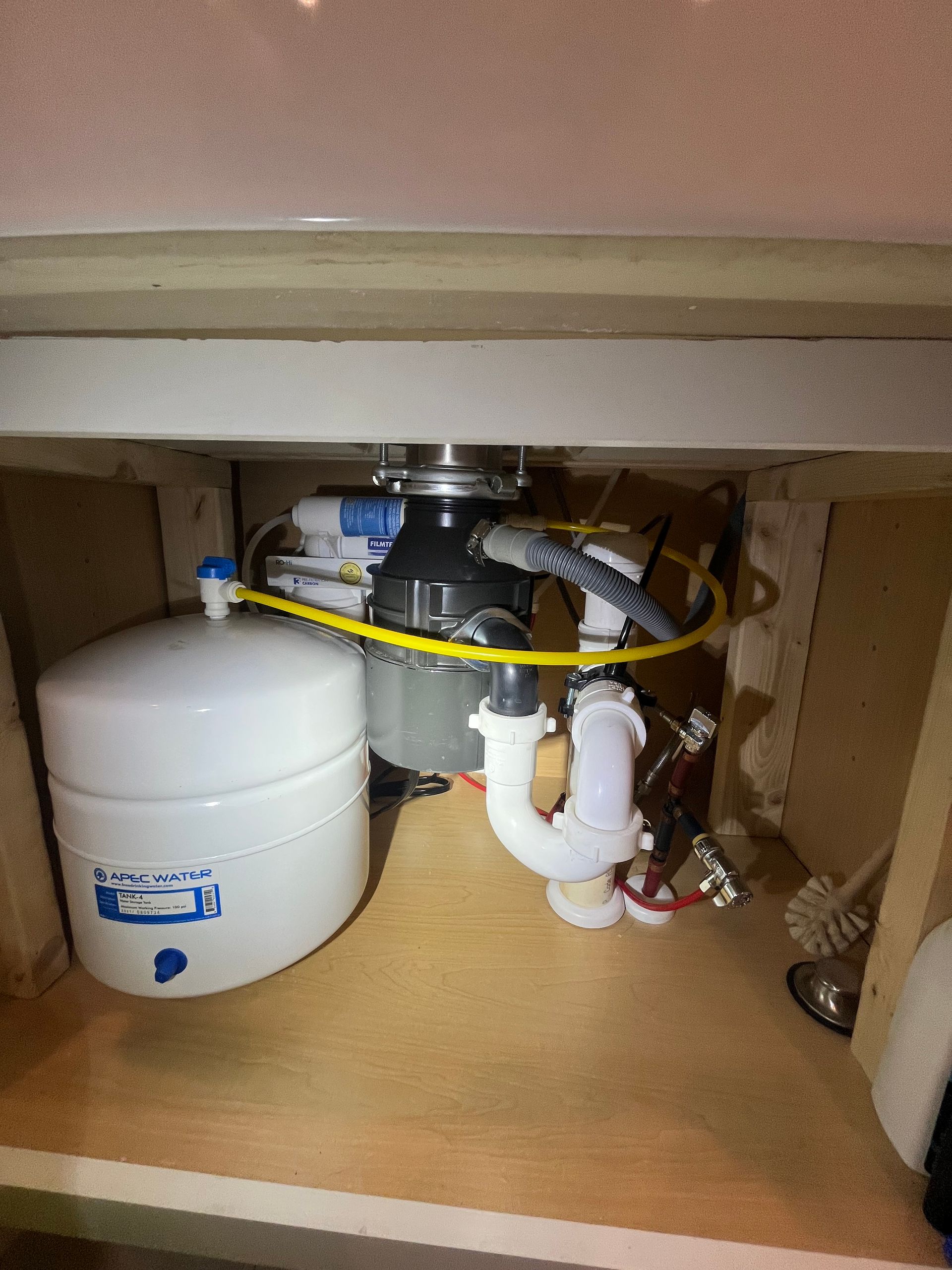 Under-sink plumbing and water filter setup with white tank, pipes, and hoses inside a cabinet