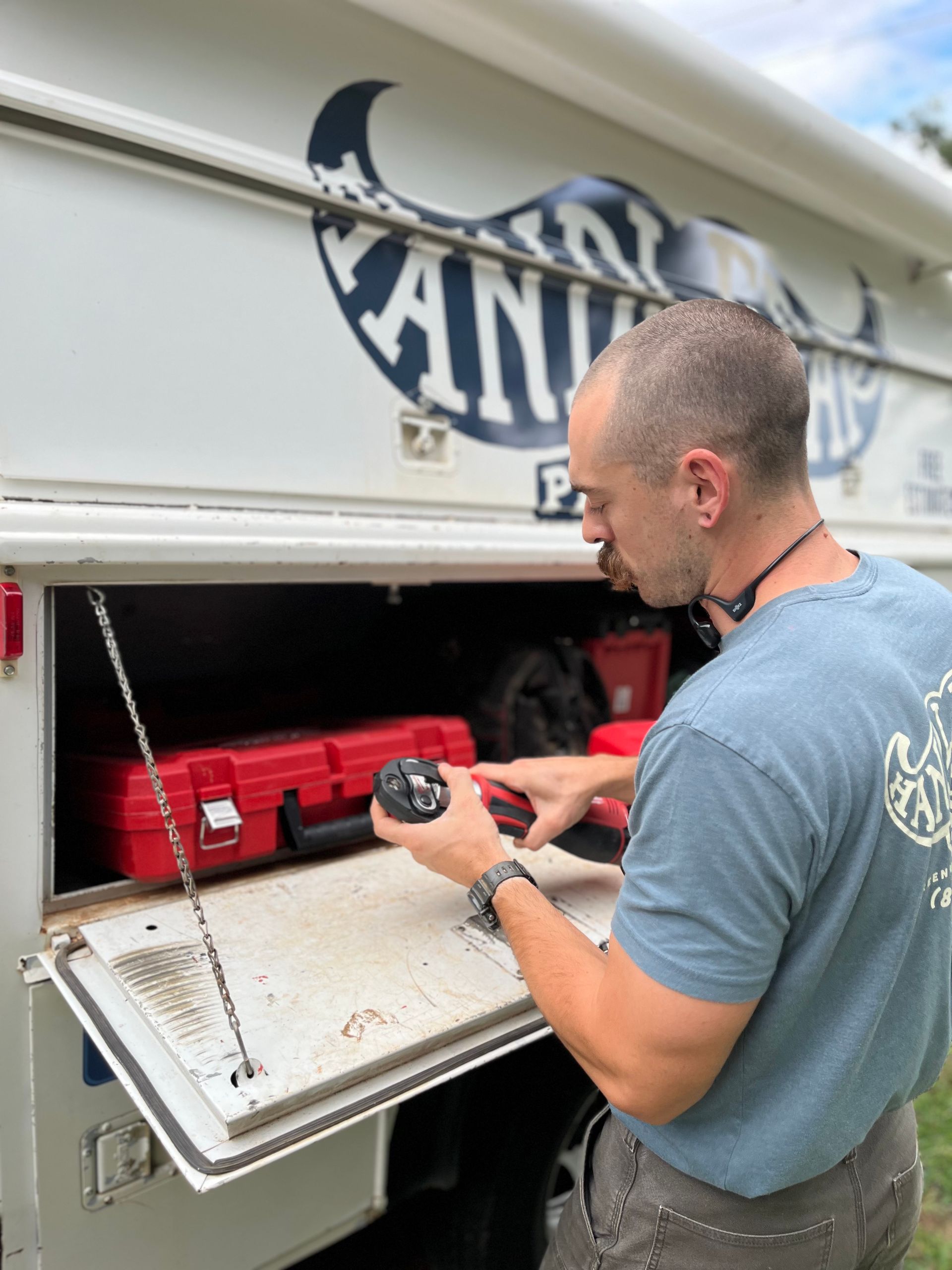 Two men stand by a white van with open doors, filled with tools. One man sits, other has arms crossed.