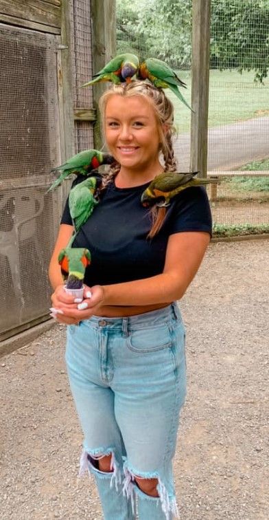 Woman holding a colorful parrot, smiling in a bird enclosure, wearing a navy shirt and ripped jeans.