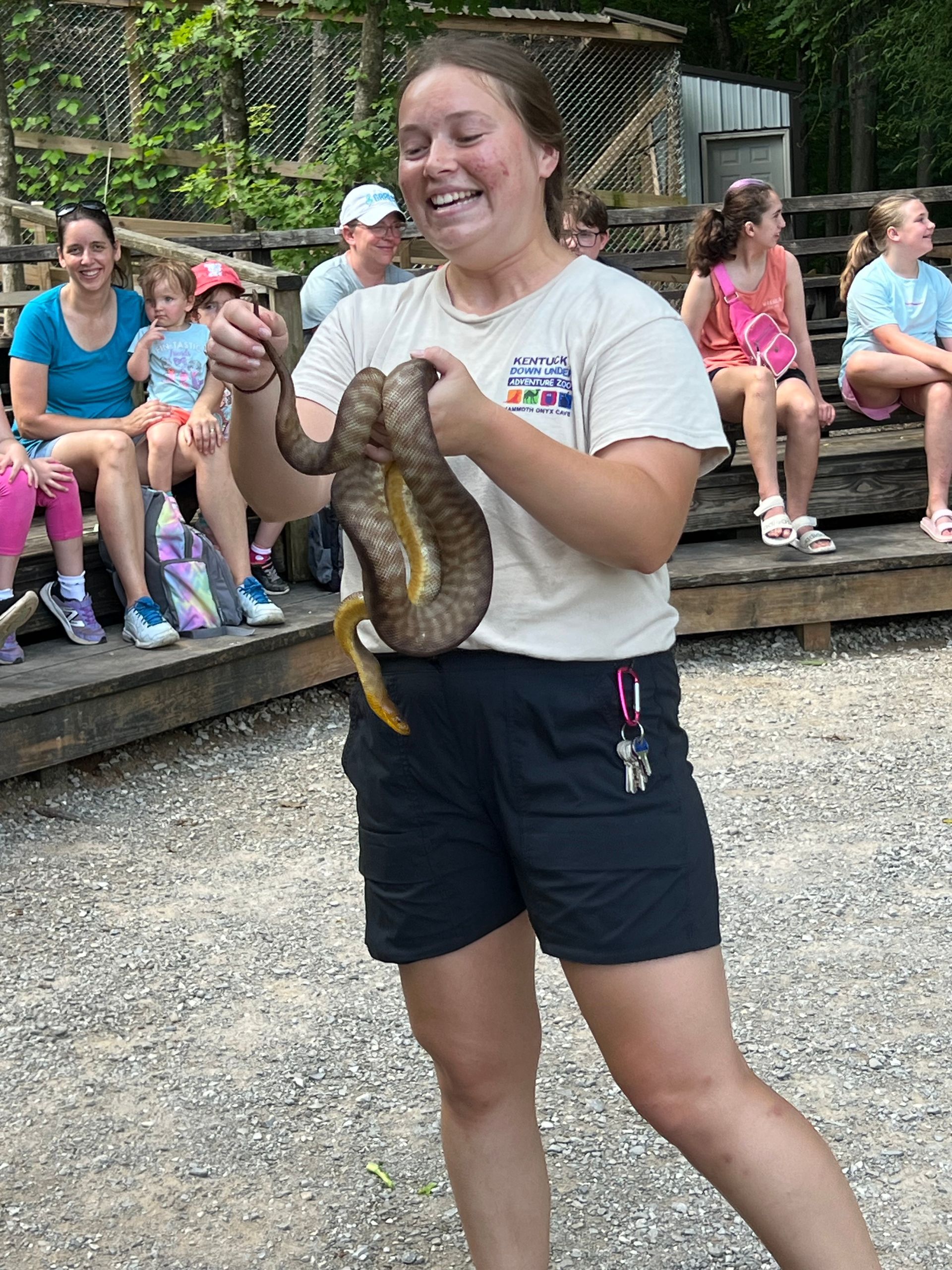 Woman holding a large snake at an outdoor wildlife show, smiling with seated spectators in the background