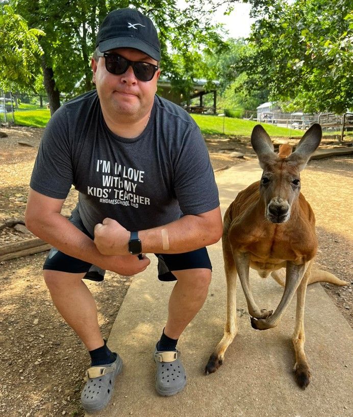 Man crouching beside a kangaroo in a leafy outdoor park, both facing the camera.