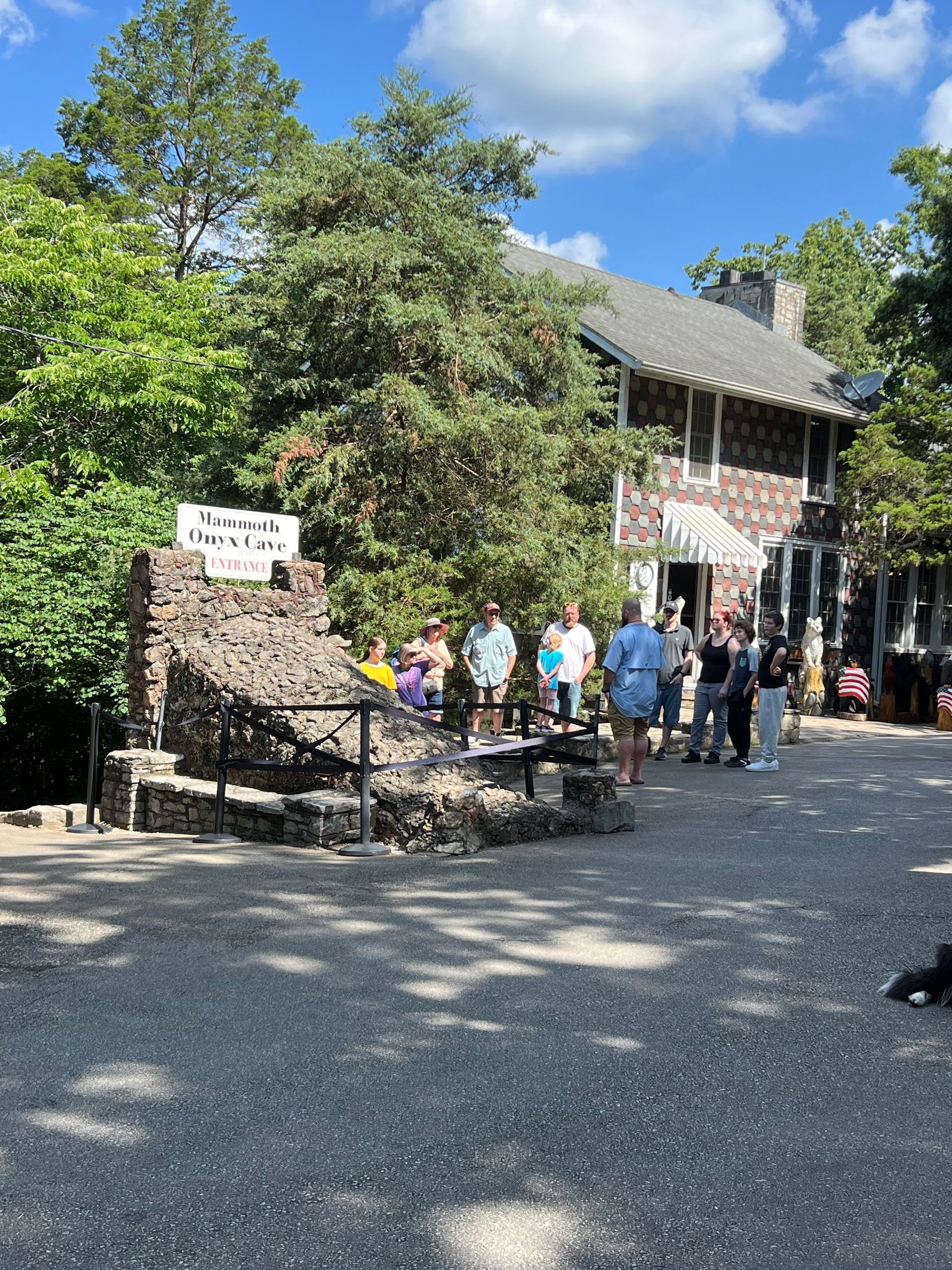 People standing outside a stone building on a sunny day, with trees and a gravel path.