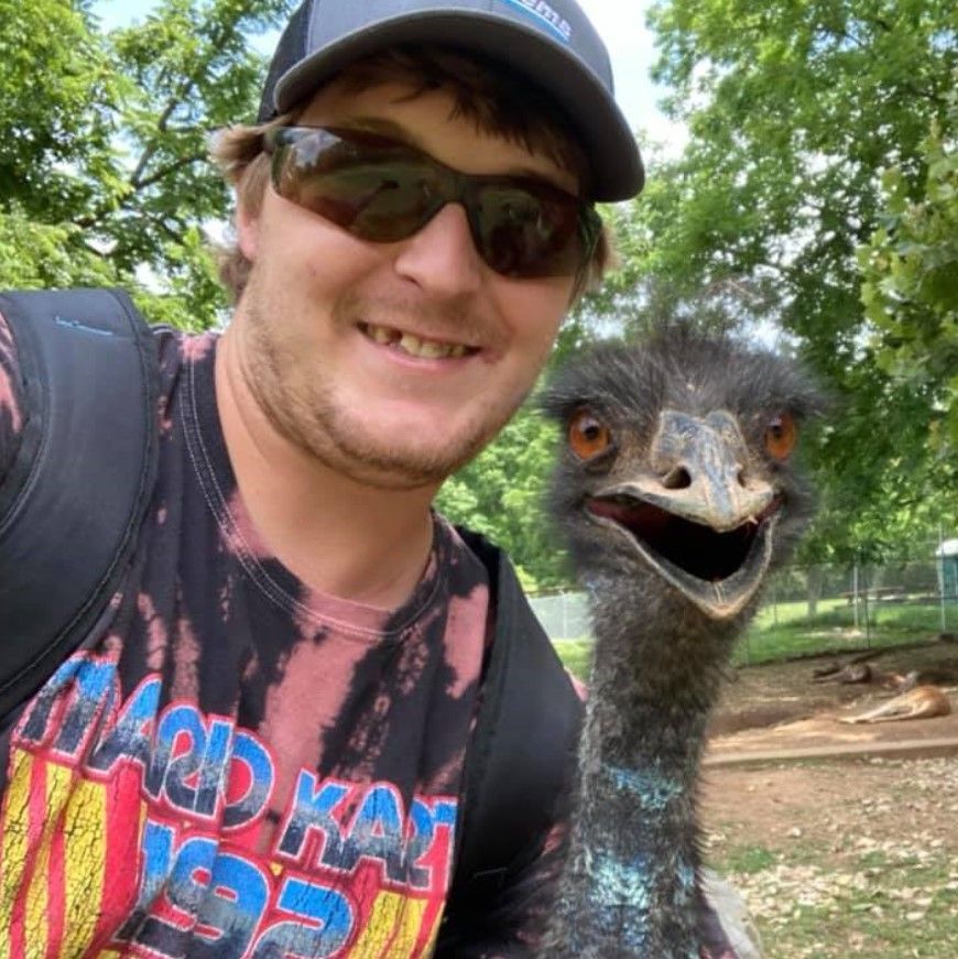 Smiling man in sunglasses posing with an emu outdoors in a wooded area