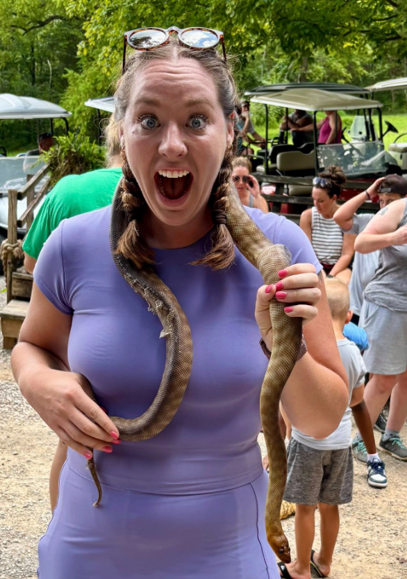 Woman in purple dress holding a snake, wide-eyed and smiling at an outdoor event