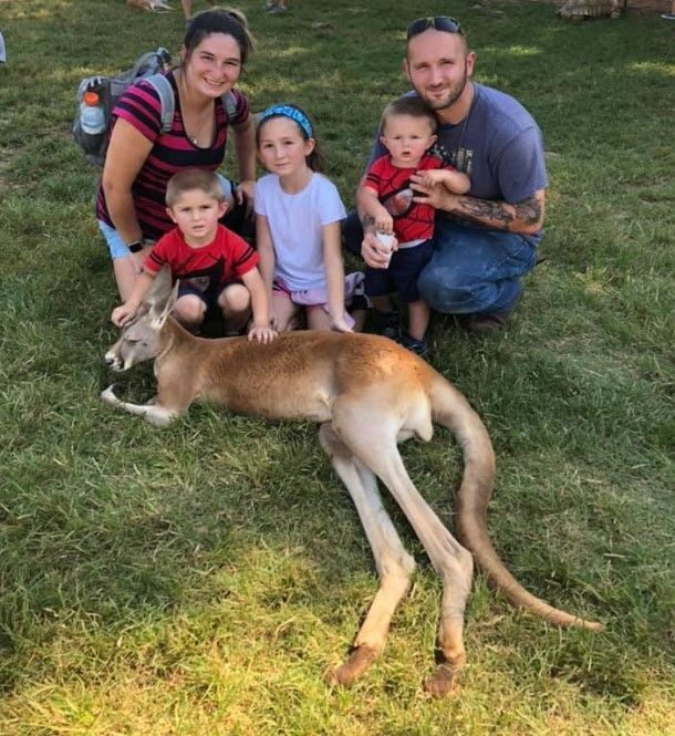Family kneeling on grass with a tan kangaroo in the foreground, smiling outdoors.