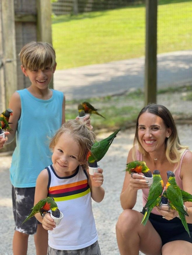 Woman and two children holding colorful parrots outdoors, smiling in a sunny park.