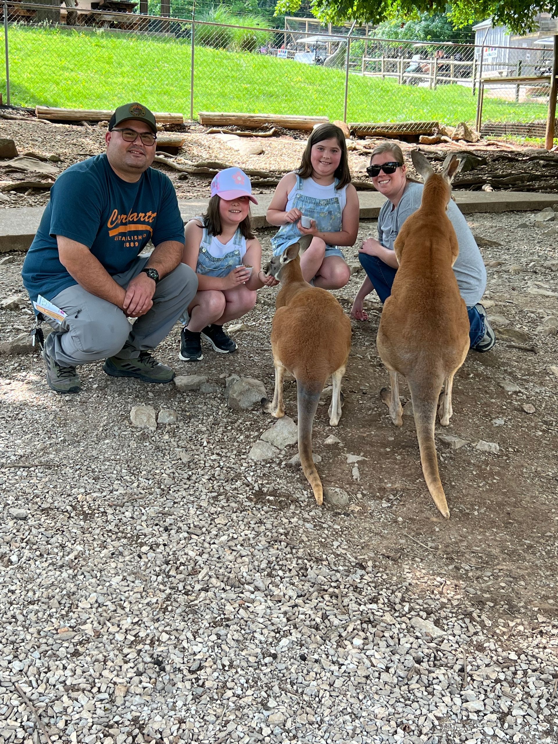 Family crouches beside two deer in a sunny outdoor park setting.