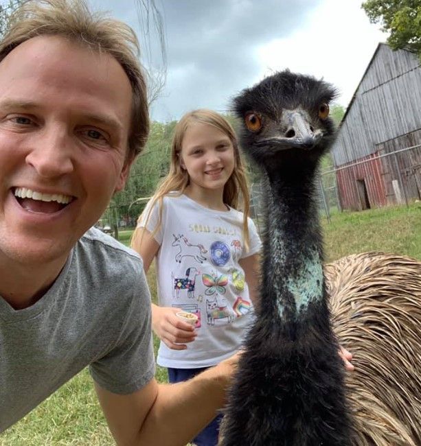 Selfie with a smiling child and an emu in a grassy yard with barns in the background