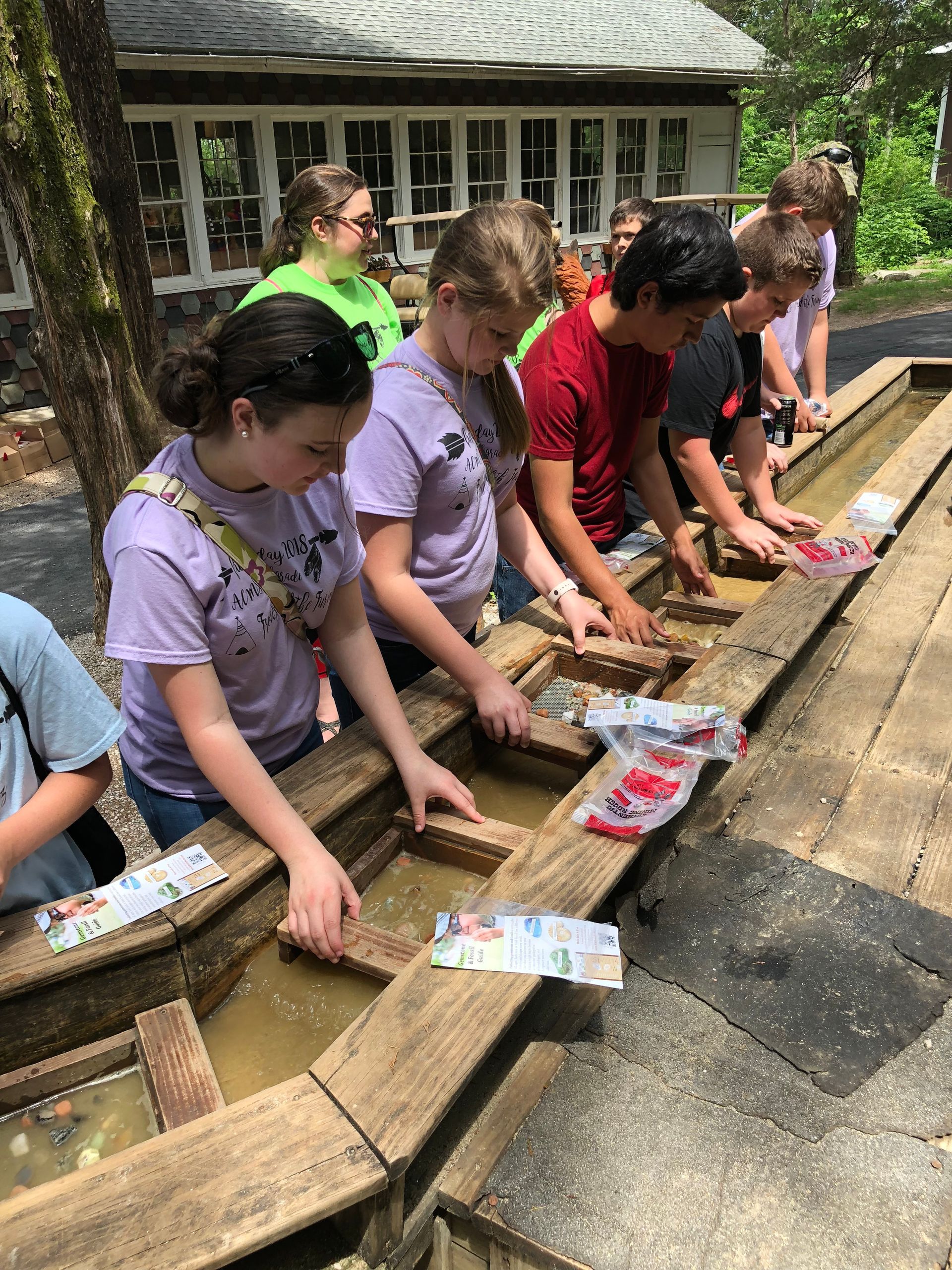 People washing paintbrushes in a wooden trough outdoors at a craft activity.