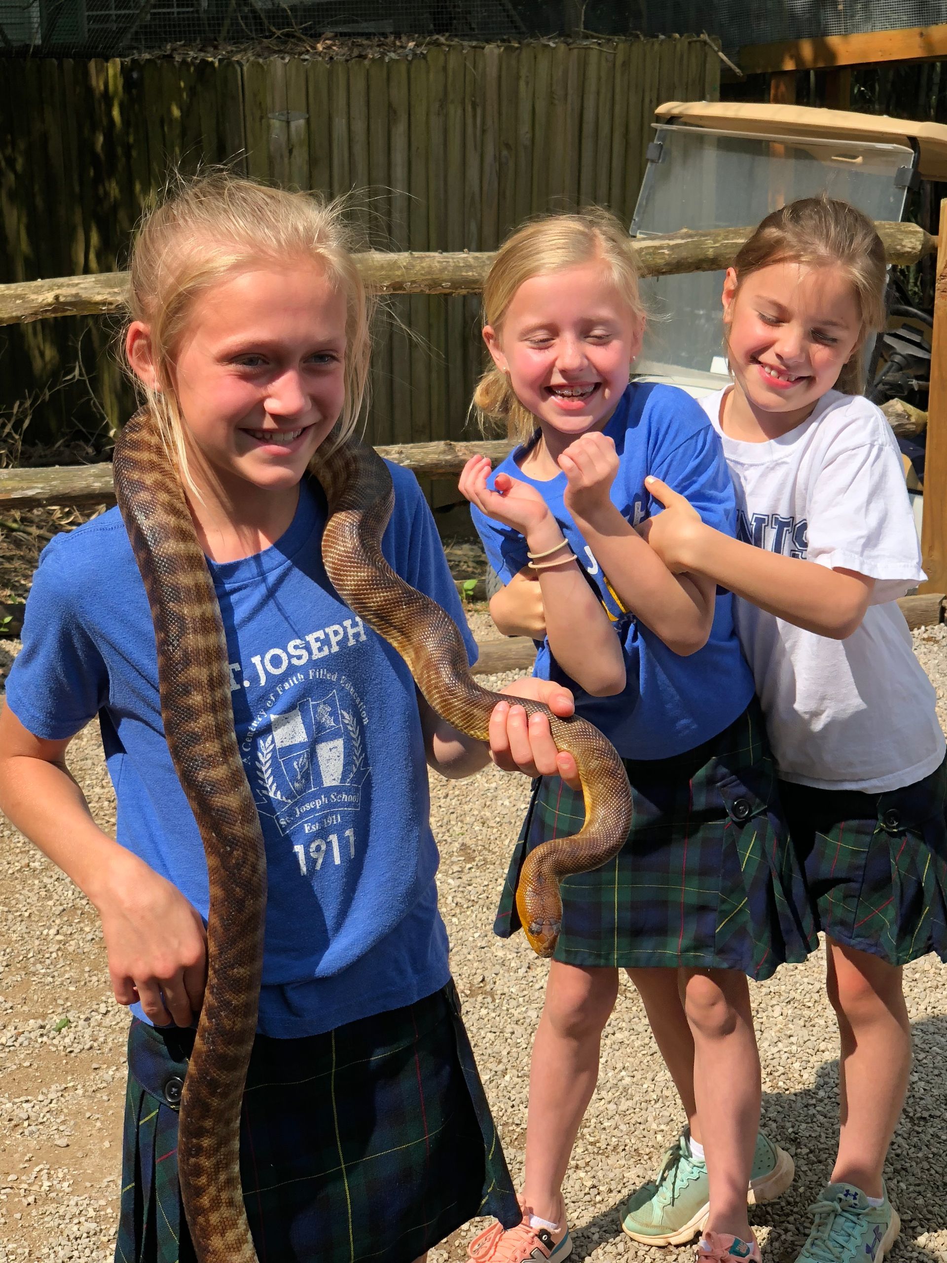 Three smiling children holding a large snake outdoors near a wooden fence
