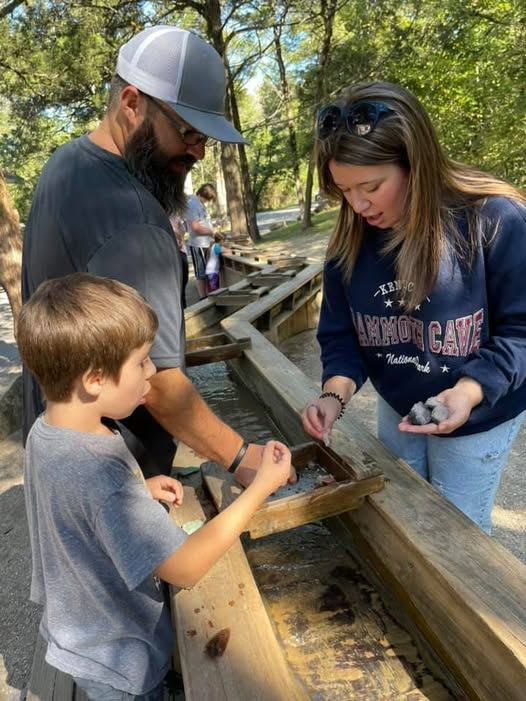 Three people examining artifacts at an outdoor wooden sluice in a park