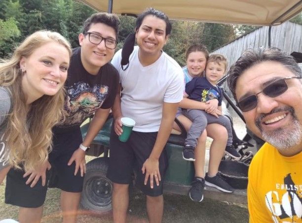 Five smiling people posing outdoors under a canopy, one holding a child and another taking the selfie.
