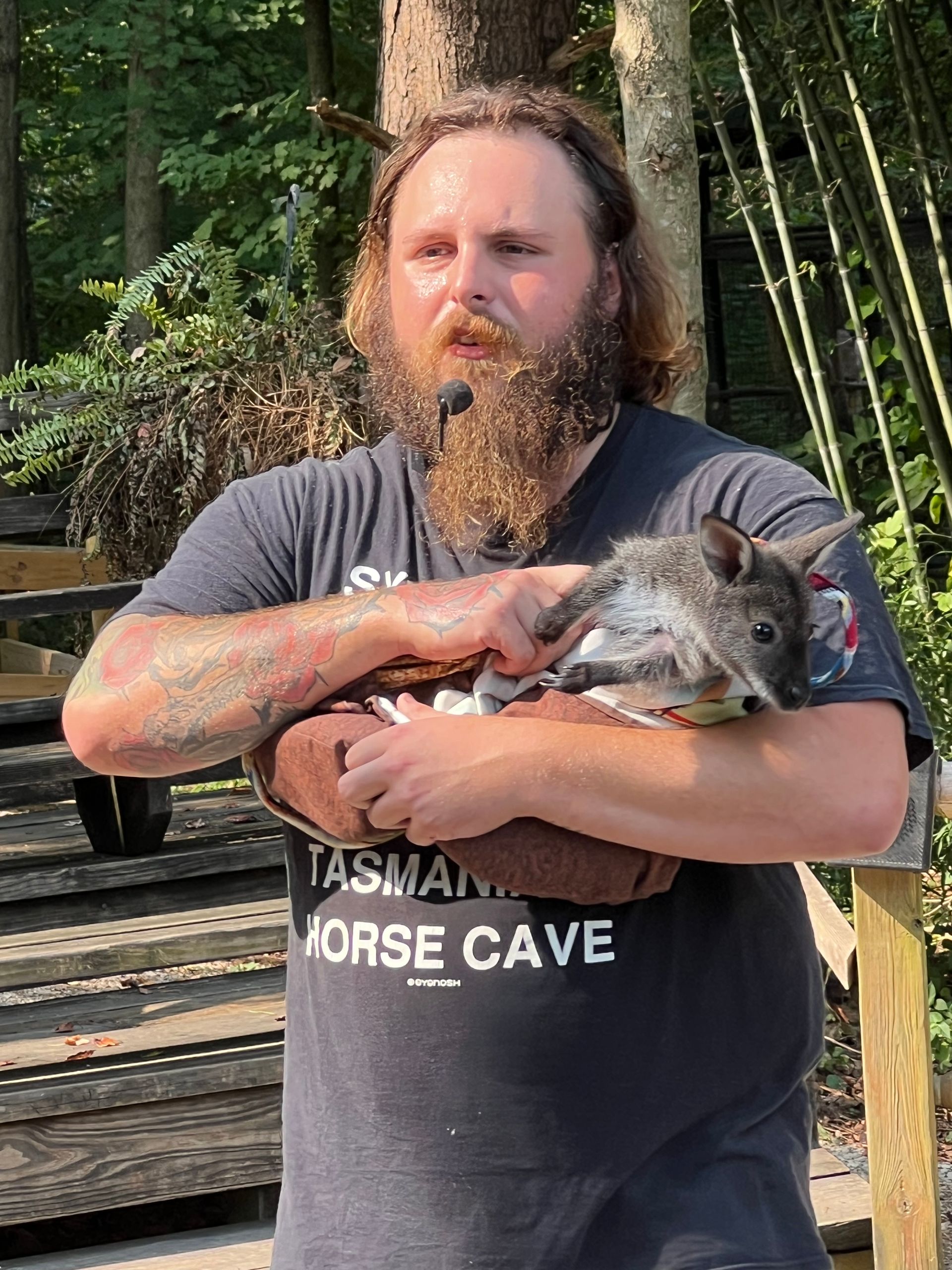 Man holding a small raccoon outdoors in front of wooden railings and trees
