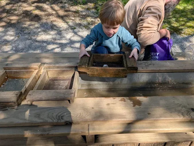 Child placing a wooden tray on a large wooden sand table outdoors with an adult nearby