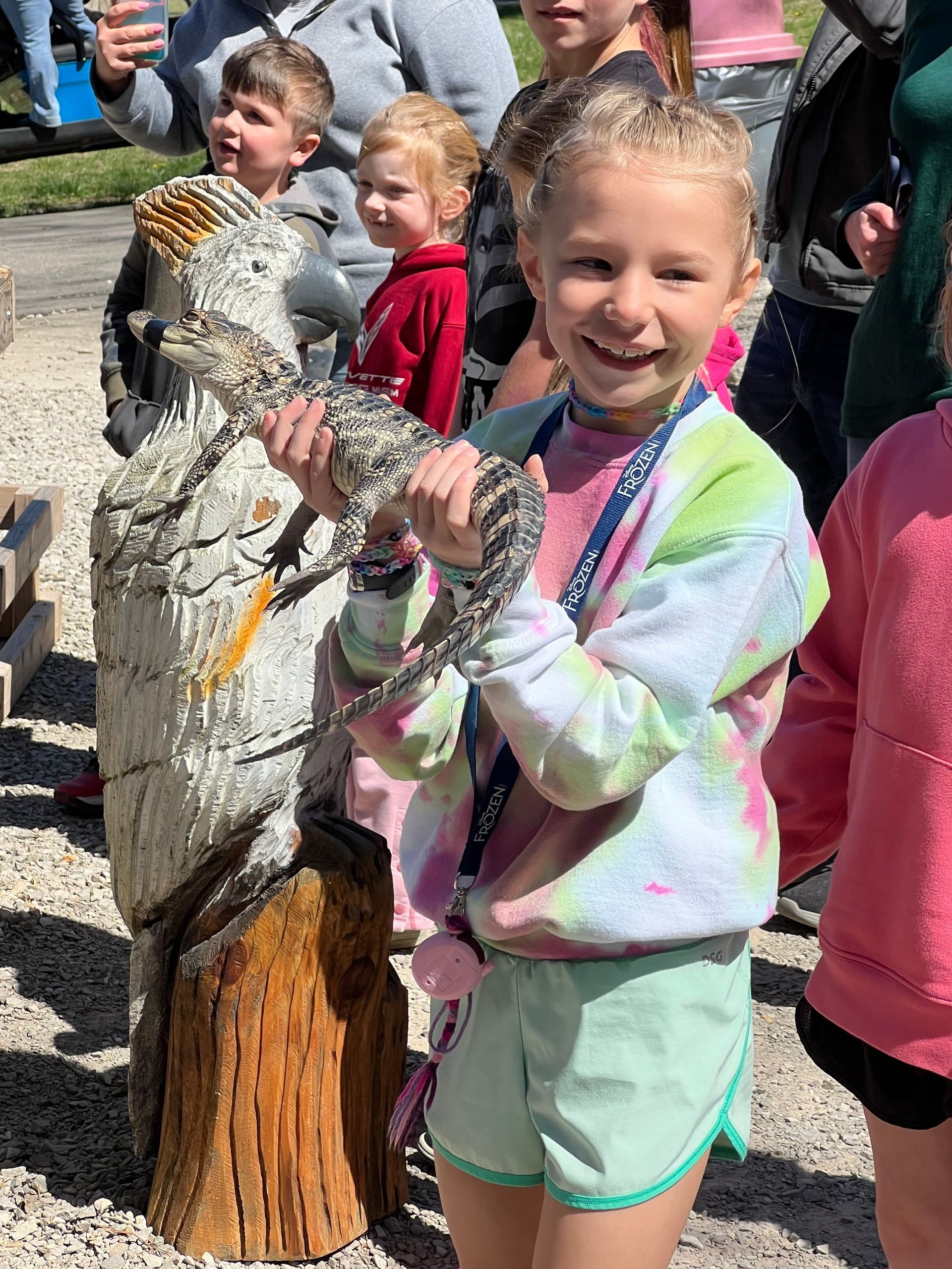 Smiling child holding a large fish on a wooden post at an outdoor event, with other children nearby