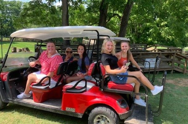 Four women riding in a red golf cart on a grassy path, smiling outdoors among trees.