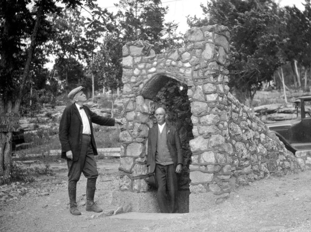 Two men standing by a stone archway in a wooded outdoor setting