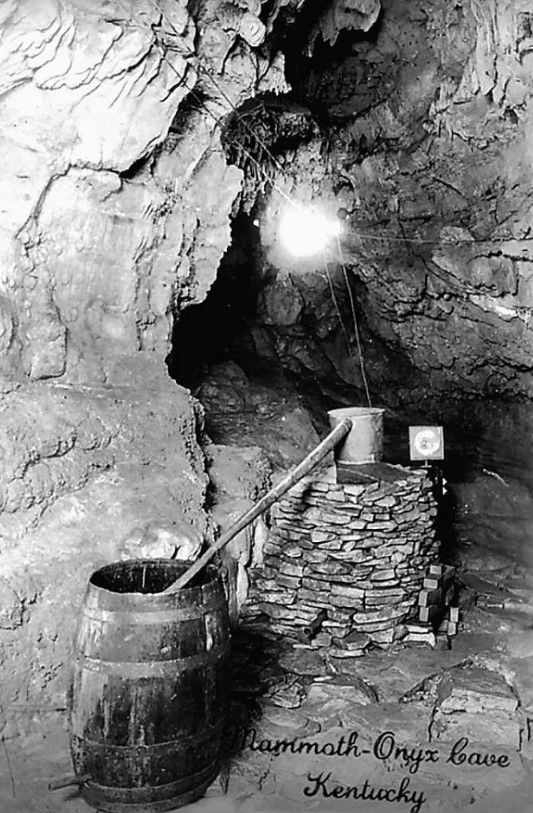Stone-lined cave passage with a barrel and a stone well under a bright lamp.