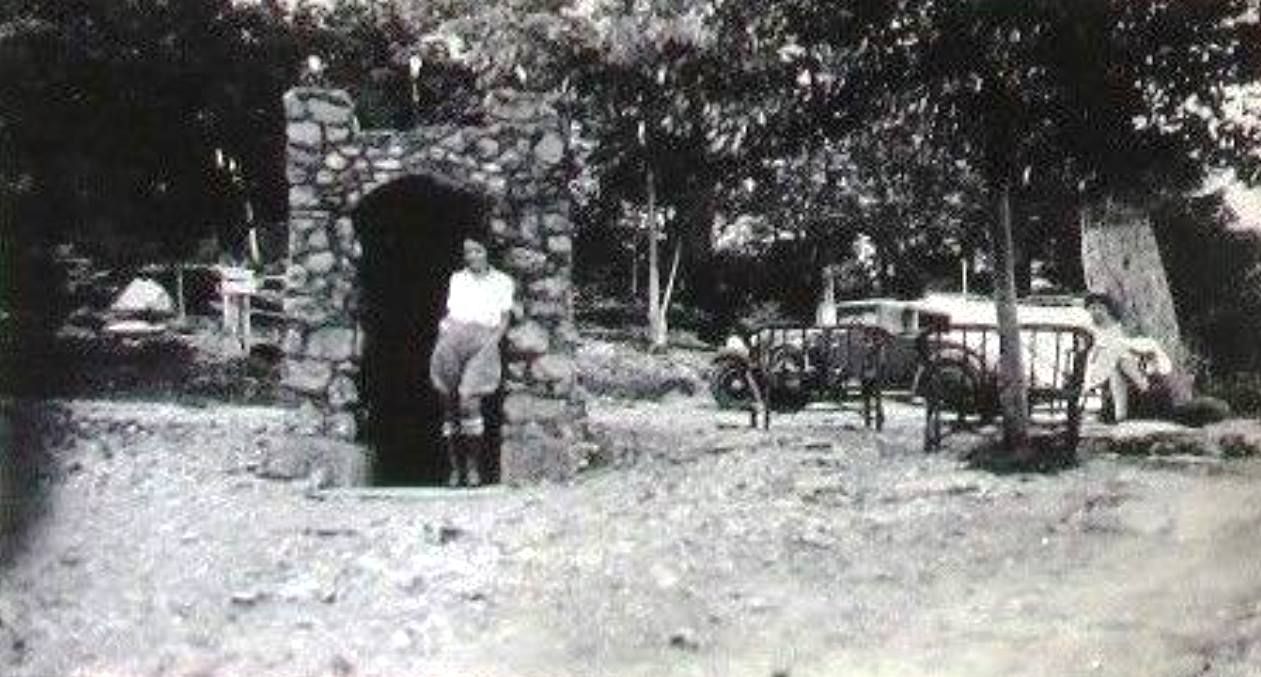 Person standing by a stone archway in a shaded outdoor courtyard with tables and chairs nearby