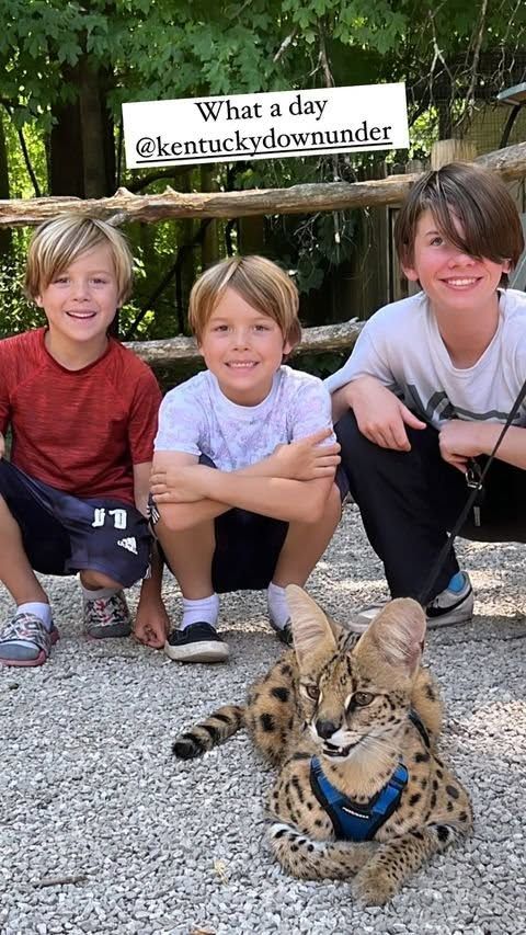 Three children crouch beside a cheetah on gravel in front of a sign at an outdoor wildlife park.