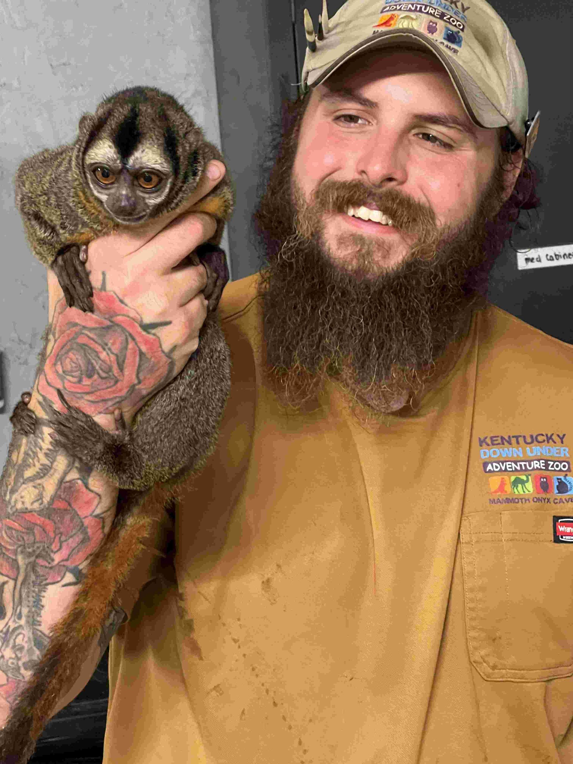 Man smiling while holding a small raccoon in a tan shirt and camo cap.