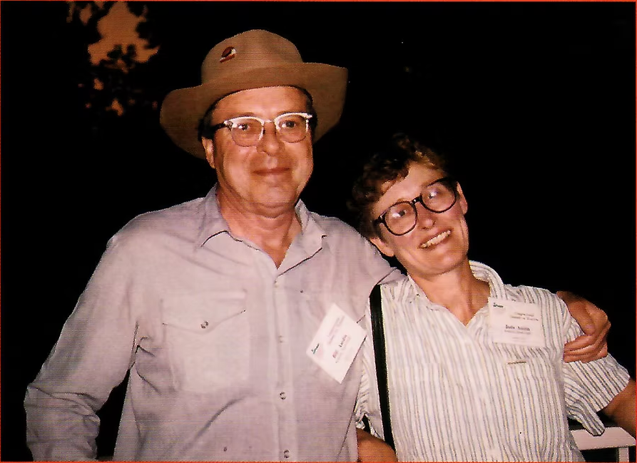 Two people smiling and posing together, wearing hats and glasses, at an indoor event.
