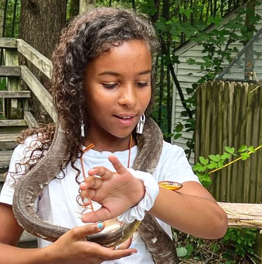 Child holding a small silver fish outdoors near a wooden fence and trees