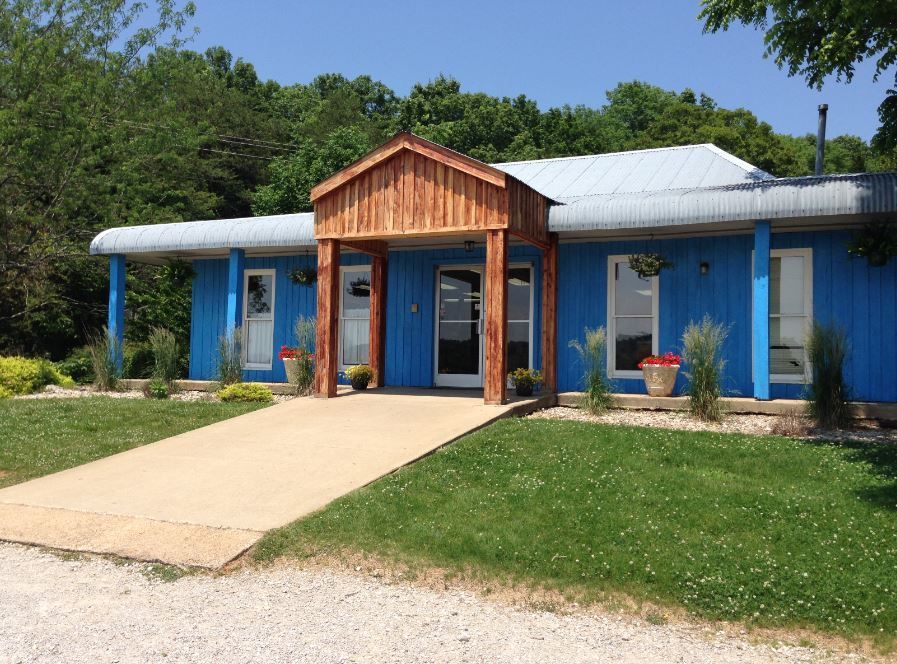 Blue building with white roof and wooden entrance, front walkway, surrounded by grass and trees.