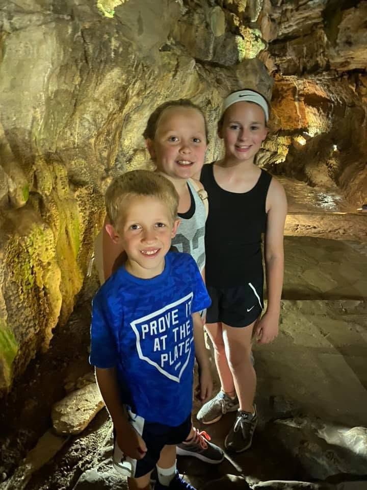Three children standing in a cave, one smiling in a blue shirt and two in black outfits.